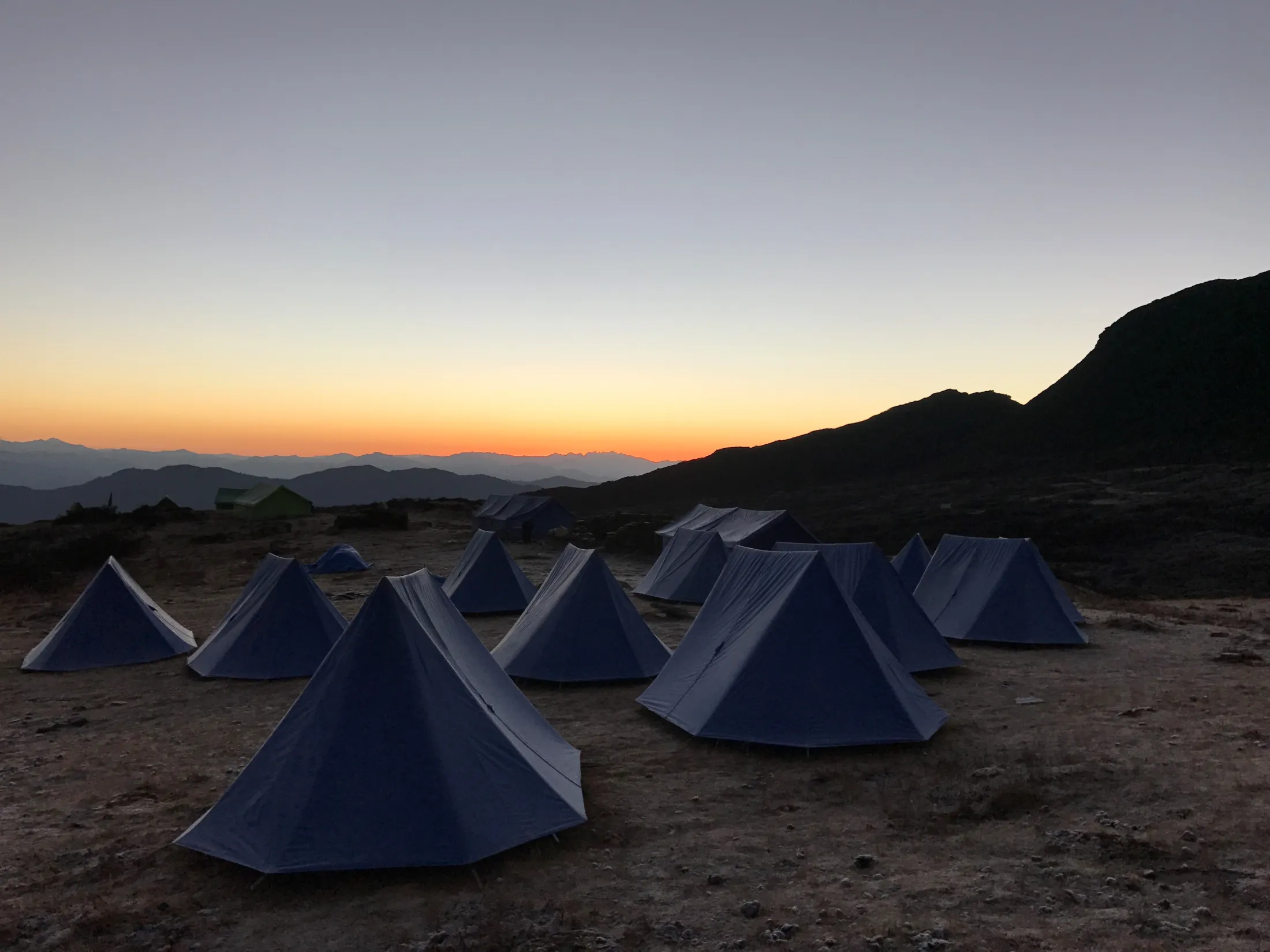 A group of blue A-frame tents set up on a high-altitude mountain campsite during a vibrant orange sunset.