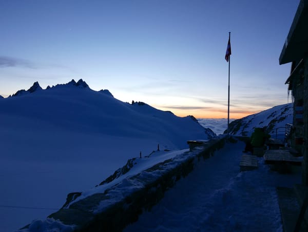 A snowy mountain landscape at dawn features a stone terrace with a Swiss flag flying on a pole against a colorful horizon.