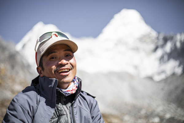A close-up portrait of a smiling man wearing a cap and sunglasses, with a beautifully blurred background of snow-capped mountain peaks against a blue sky.