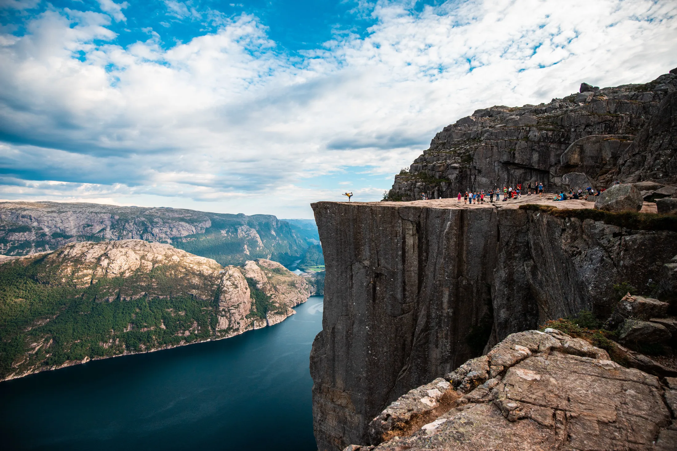 Hikers standing on top of the steep cliff Preikestolen overlooking a deep fjord in Norway under a cloudy sky.