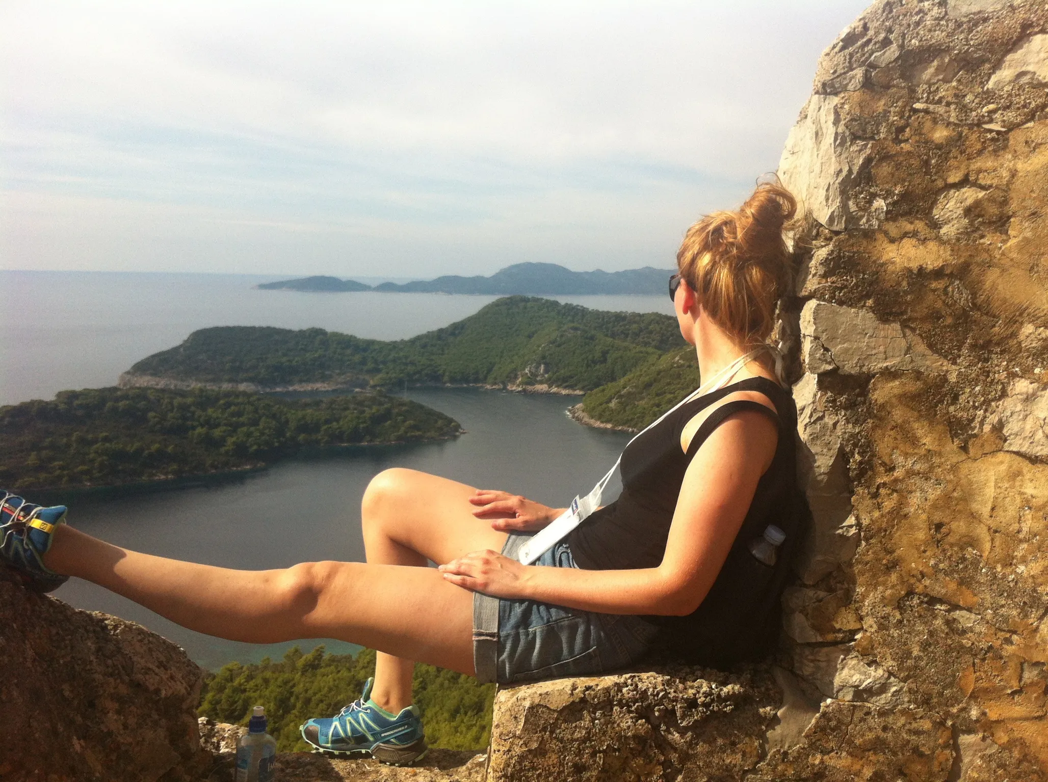 A woman relaxes on a stone ledge, looking out over a scenic vista of forested islands and blue water.