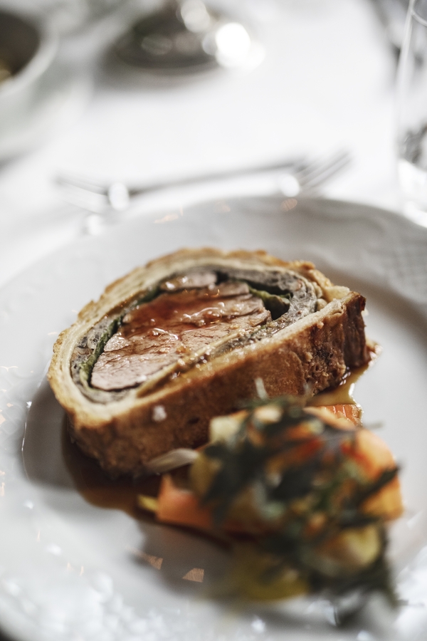 A close-up shot of a slice of Beef Wellington with pastry crust and pink meat, served on a white plate with side vegetables and sauce.