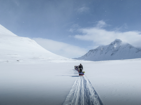 A person on a snowmobile driving across a vast snowy landscape in Svalbard, with mountains in the background under a partly cloudy sky.