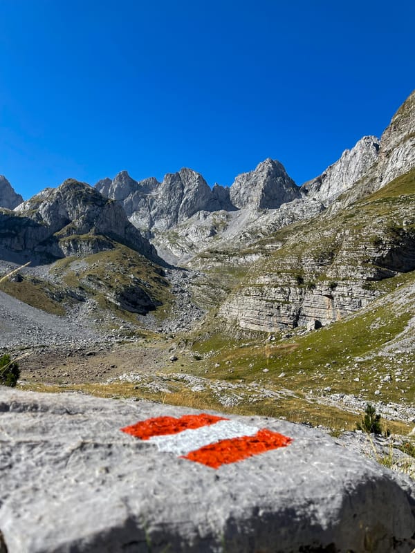 A red and white trail marker painted on a rock with a mountainous landscape in the background under a blue sky.