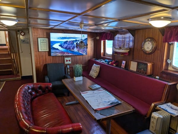 A cozy wood-paneled ship lounge with red leather sofas, a table, and a wall-mounted TV.