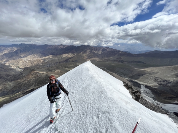 A mountaineer equipped with a backpack and a trekking pole navigates a steep, sharp snow ridge, with a vast expanse of barren mountain valleys stretching below a cloudy sky.