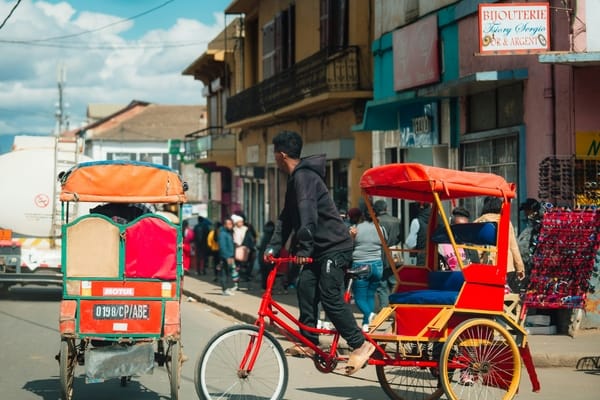 A young man dressed in dark clothes rides a bright red cycle rickshaw down a busy city street flanked by colorful storefronts.