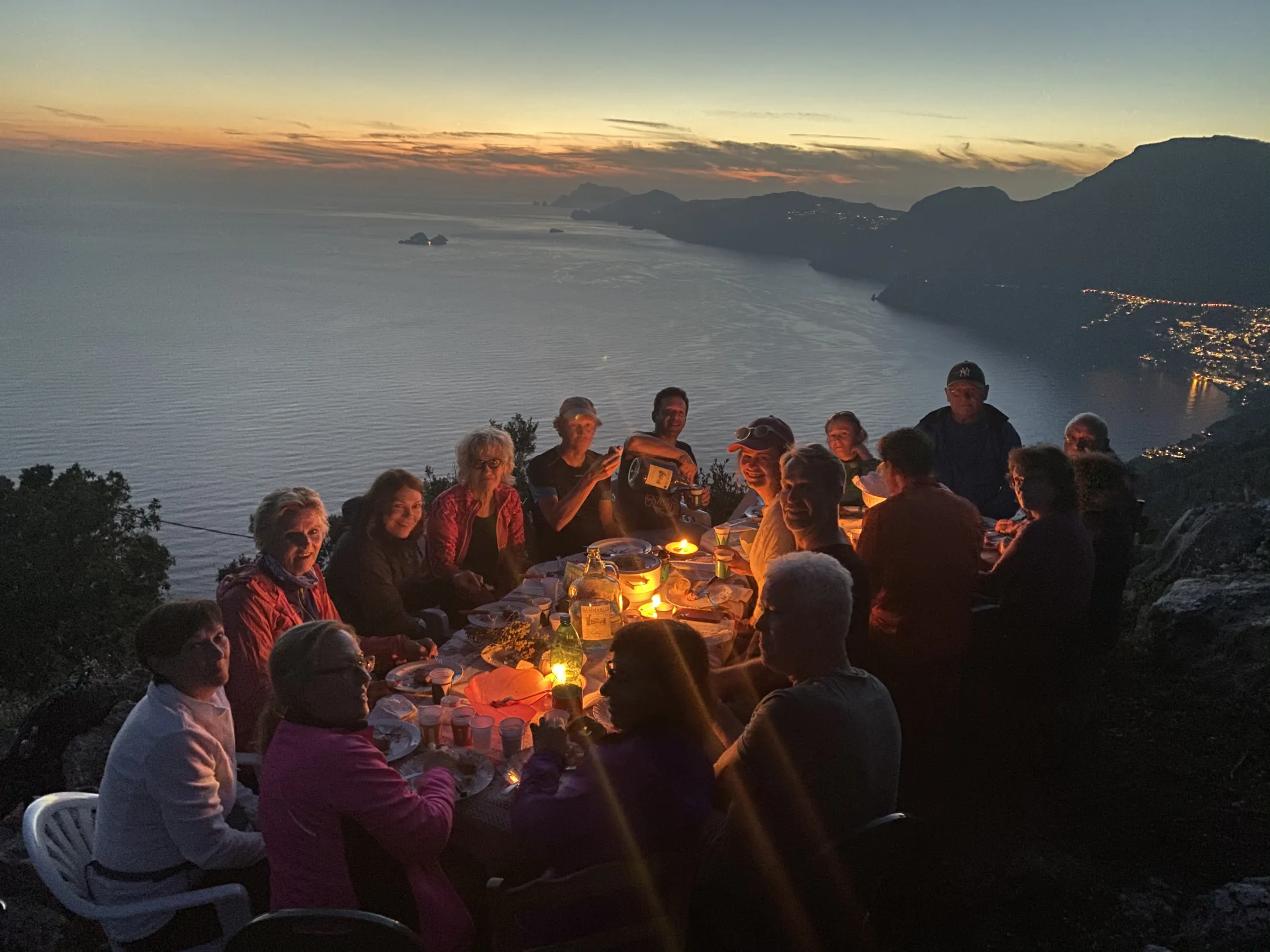 A large group of people dining outdoors at a candlelit table overlooking the sea at sunset.