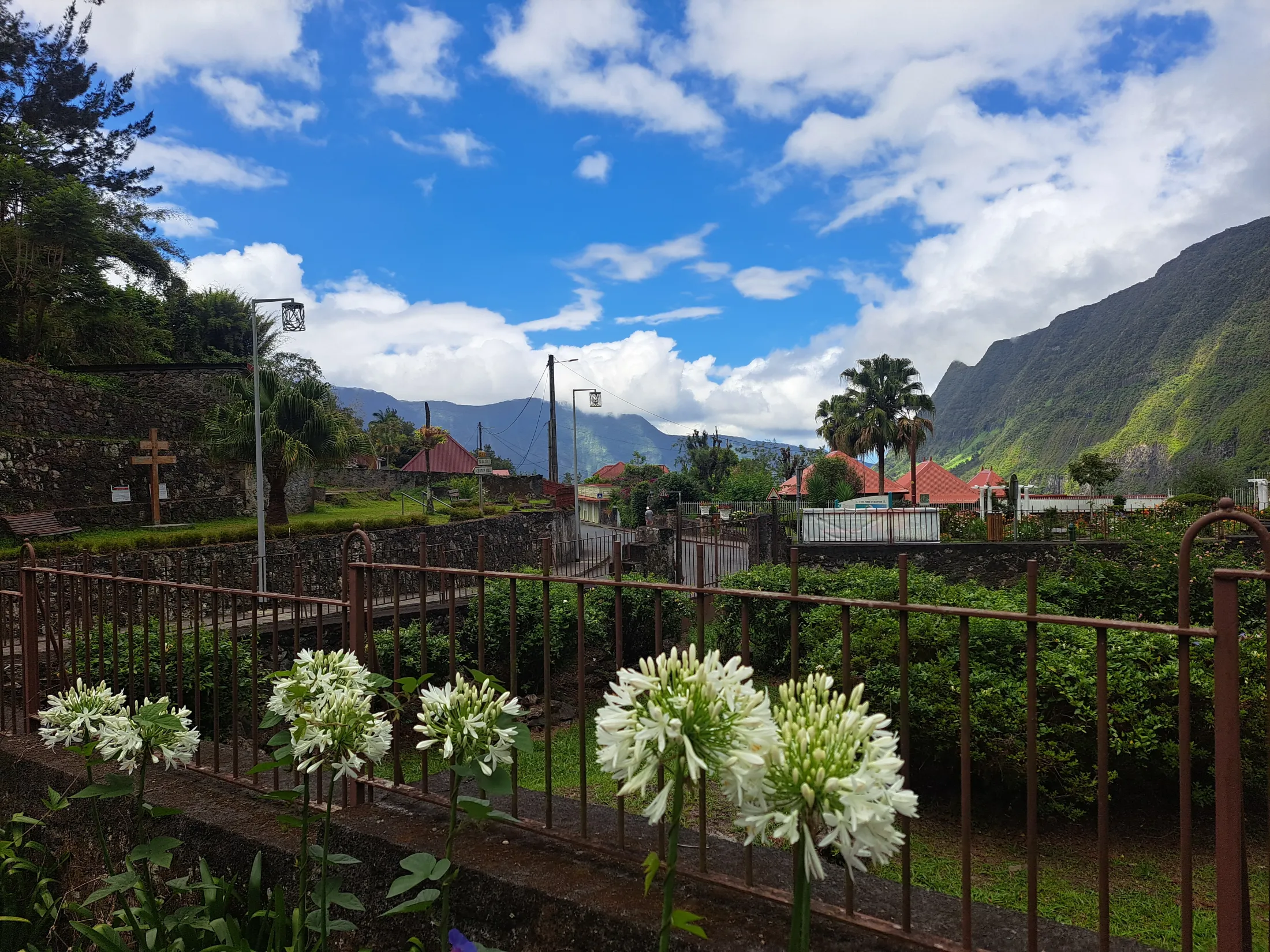 White flowers bloom behind a metal fence in the foreground of a charming mountain village with red-roofed buildings and towering green peaks under a cloudy blue sky.