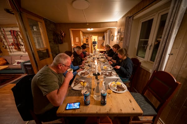 A group sitting around a long wooden dining table, eating and drinking in a cabin interior.