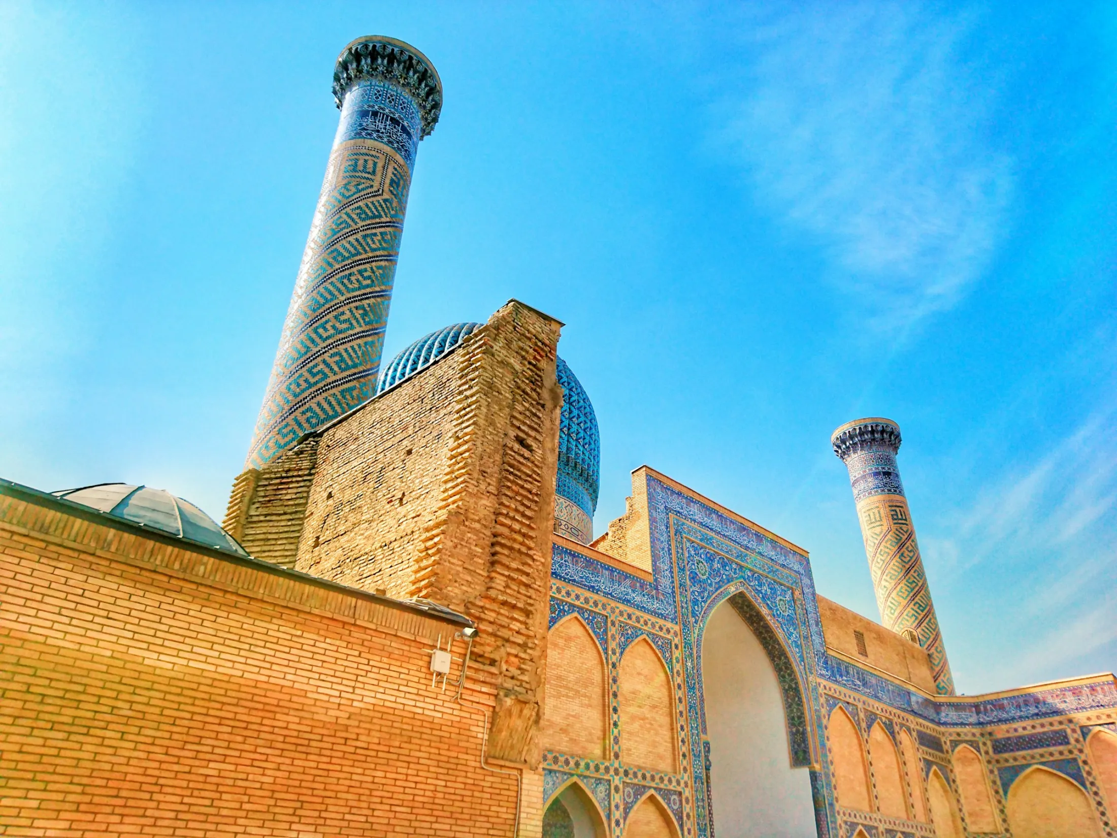 The low-angle shot features a historic brick mosque structure adorned with intricate blue mosaic tiles and two towering minarets under a clear sky.