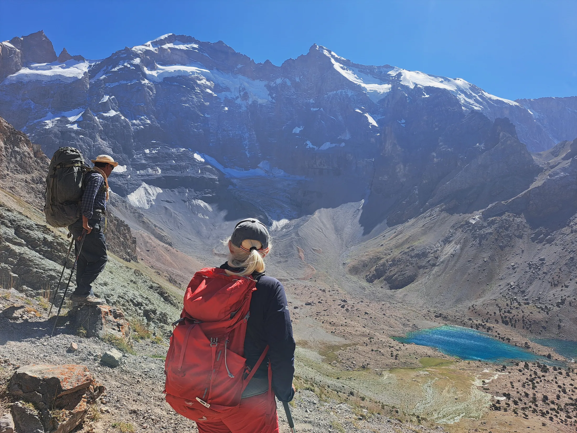Two hikers with large backpacks look out over a high mountain valley featuring a bright blue alpine lake and massive, snow-covered peaks under a clear sky.