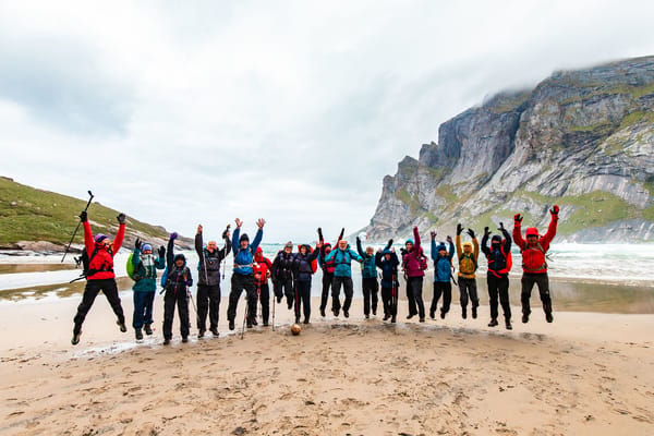 A large group of people jumping for joy on a sandy beach with mountains in the background.