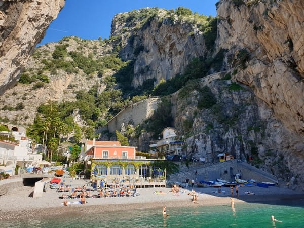 A beach with sunbeds and buildings nestled between dramatic rocky cliffs, with turquoise water in the foreground.