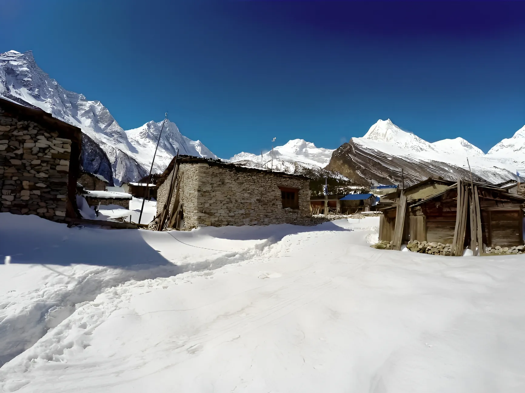 Rustic stone houses sit nestled in deep snow within a remote village, surrounded by majestic, snow-covered mountain peaks under a clear blue sky.