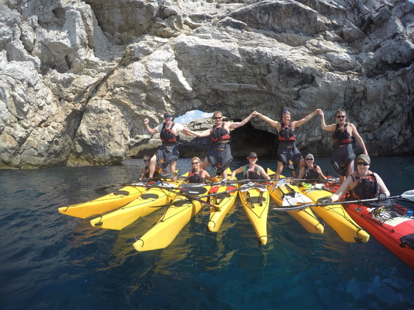 A group of kayakers celebrate together on calm blue water, with several people standing up and holding hands across their yellow and red kayaks, positioned in front of a massive rocky cliff featuring a natural stone arch.