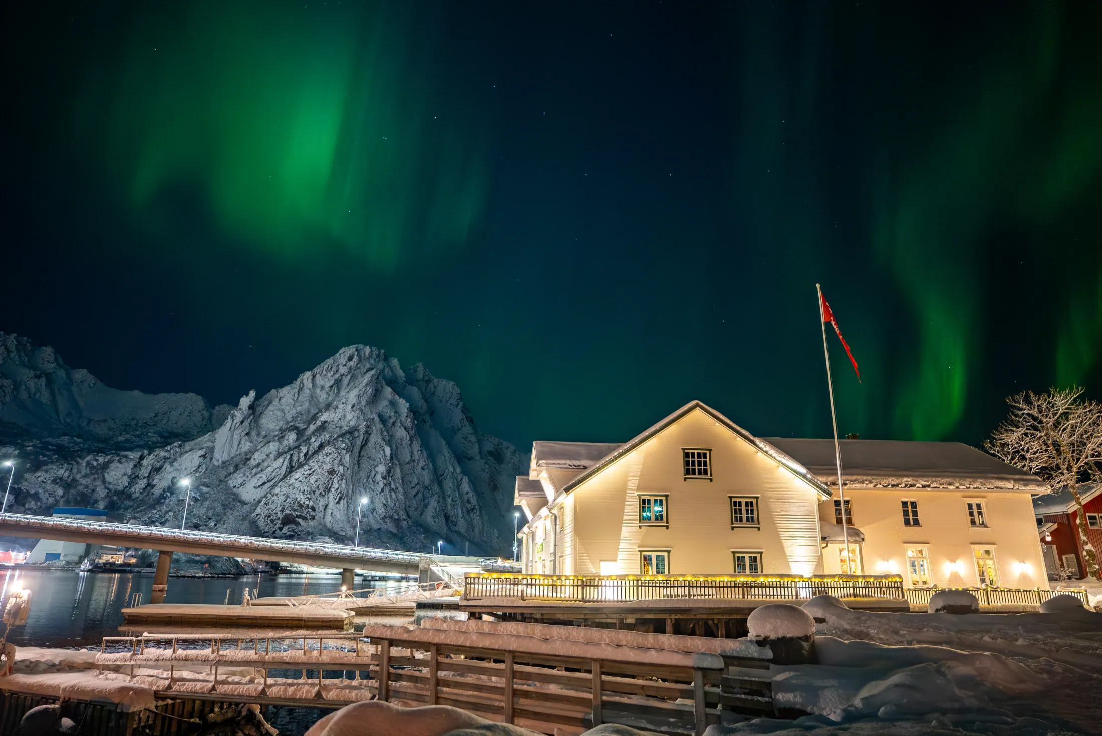 Northern lights in a dark night sky, with snow-covered ground in the foreground, a white wooden house, and an alpine mountain in the background.