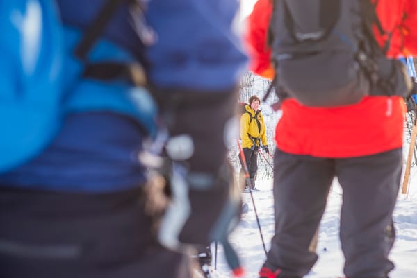 Hikers standing in the snow, viewed from behind with out-of-focus figures in the foreground.