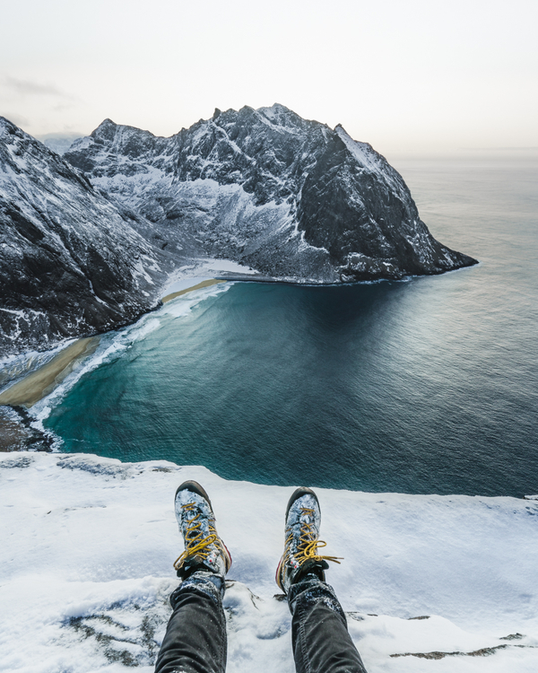 Feet hanging over the ledge of a mountain looking down on the beach and ocean in the snow