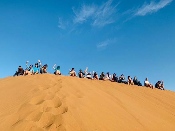 A group of people sitting on top of a sand dune with blue skies