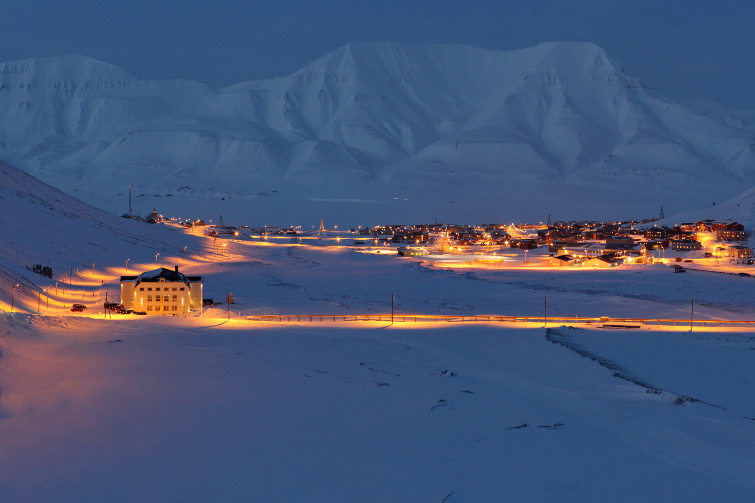 Snowy landscape and mountains with a street and buildings with some street lights