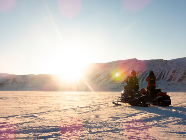 Two people on snowmobiles in a low sun with mountains
