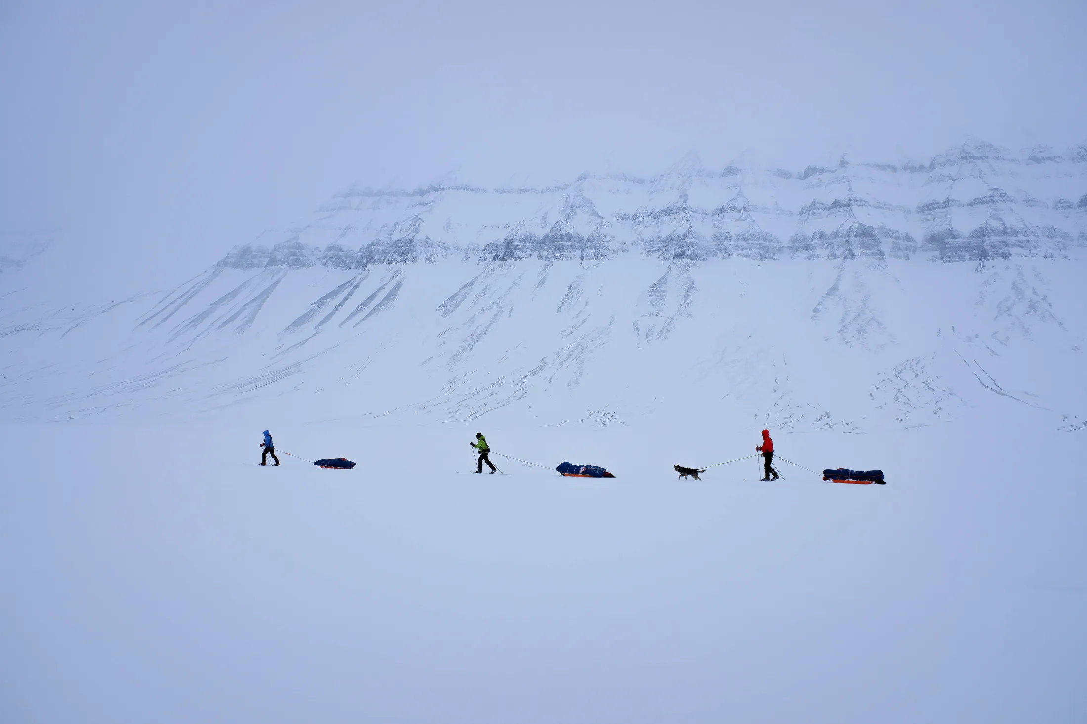 Tre skiløpere trekker lastede pulker over en vid snøslette med en hund ved siden av seg, på bakgrunn av et enormt, snødekket fjell med tydelige horisontale berglag.