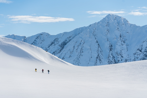 Tre turgåere i et stort, snødekket landskap med høye fjell i bakgrunnen under en blå himmel.