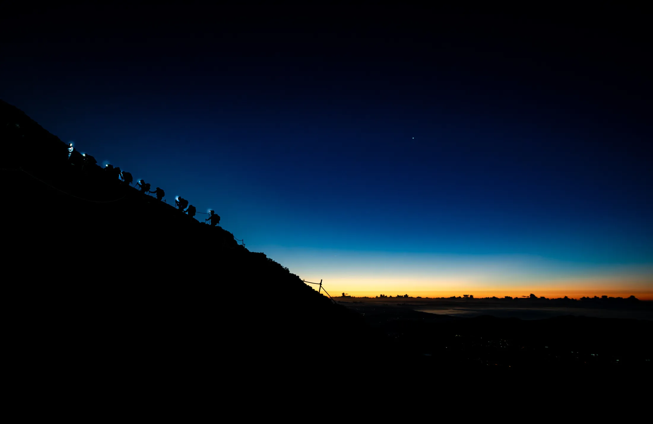 Silhuetter av turgåere med hodelykter klatrer opp en bratt fjellside under en mørkeblå nattehimmel som glir over i en fargerik, oransje soloppgangshorisont.