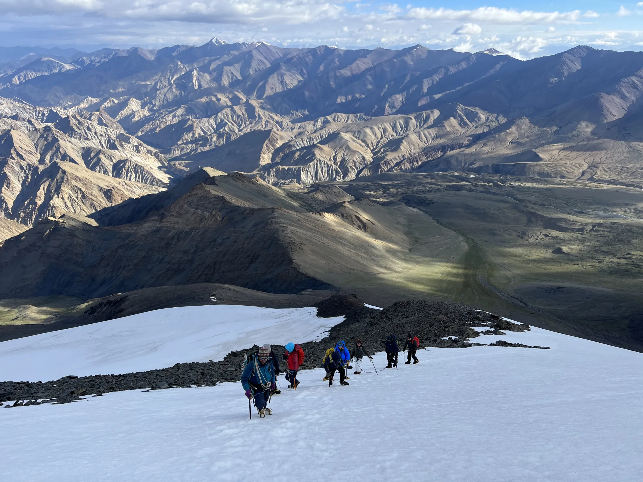 Et lag med fjellklatrere beveger seg over en bratt, snødekt skråning, med et enormt landskap av kuperte, golde fjellrygger og dype daler som strekker seg ut langt under dem under en mild himmel.