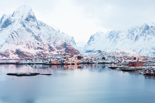 Et vinterlandskap med en kystby omgitt av majestetiske snødekte fjell, speilblankt i vannet.