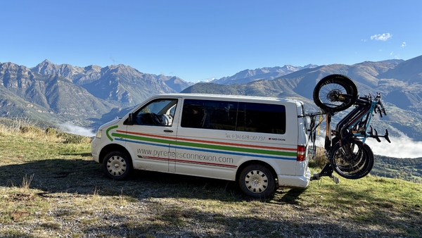 En hvit bobil parkert på en fjellhylle med sykler festet bak, med utsikt over en dyp dal og fjerne fjelltopper under en klar blå himmel.