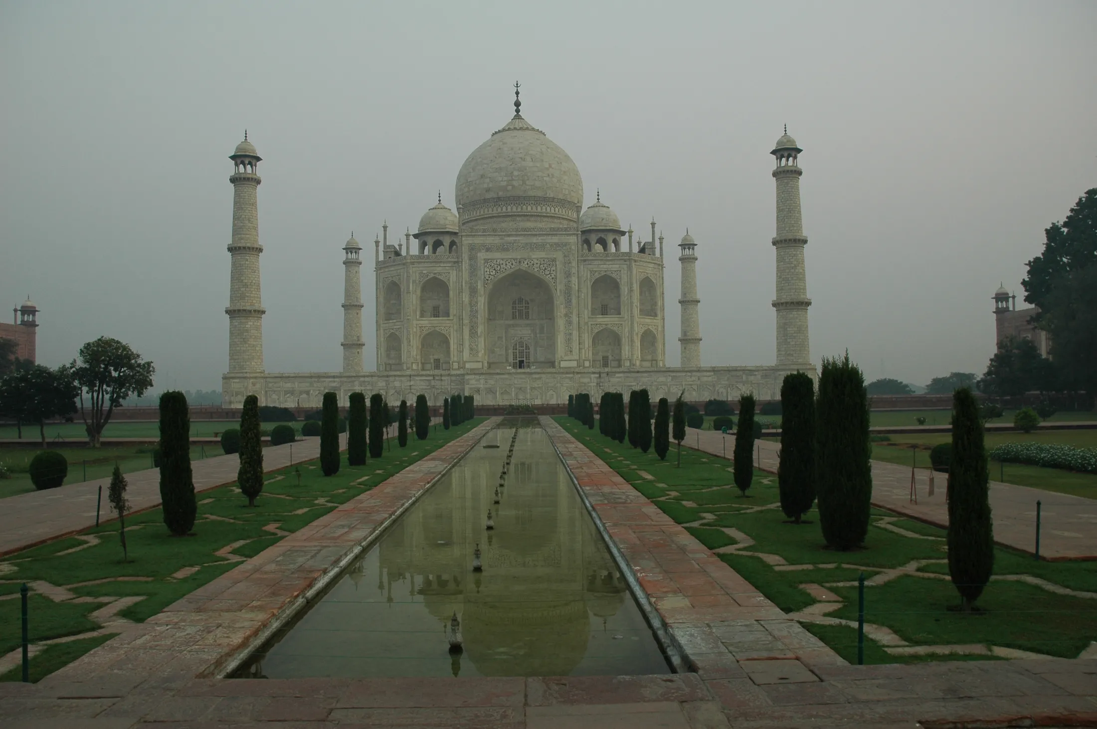 Det ikoniske, hvite marmorpalasset Taj Mahal står majestetisk i enden av et langt speilbasseng, omkranset av symmetriske gressplener og frisierte trær under en disig, overskyet himmel.