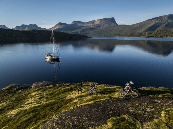 Tre terrengsyklister dytter sykkelen opp en gressbevokst skråning med utsikt over en rolig fjord med en seilbåt og fjell i bakgrunnen.