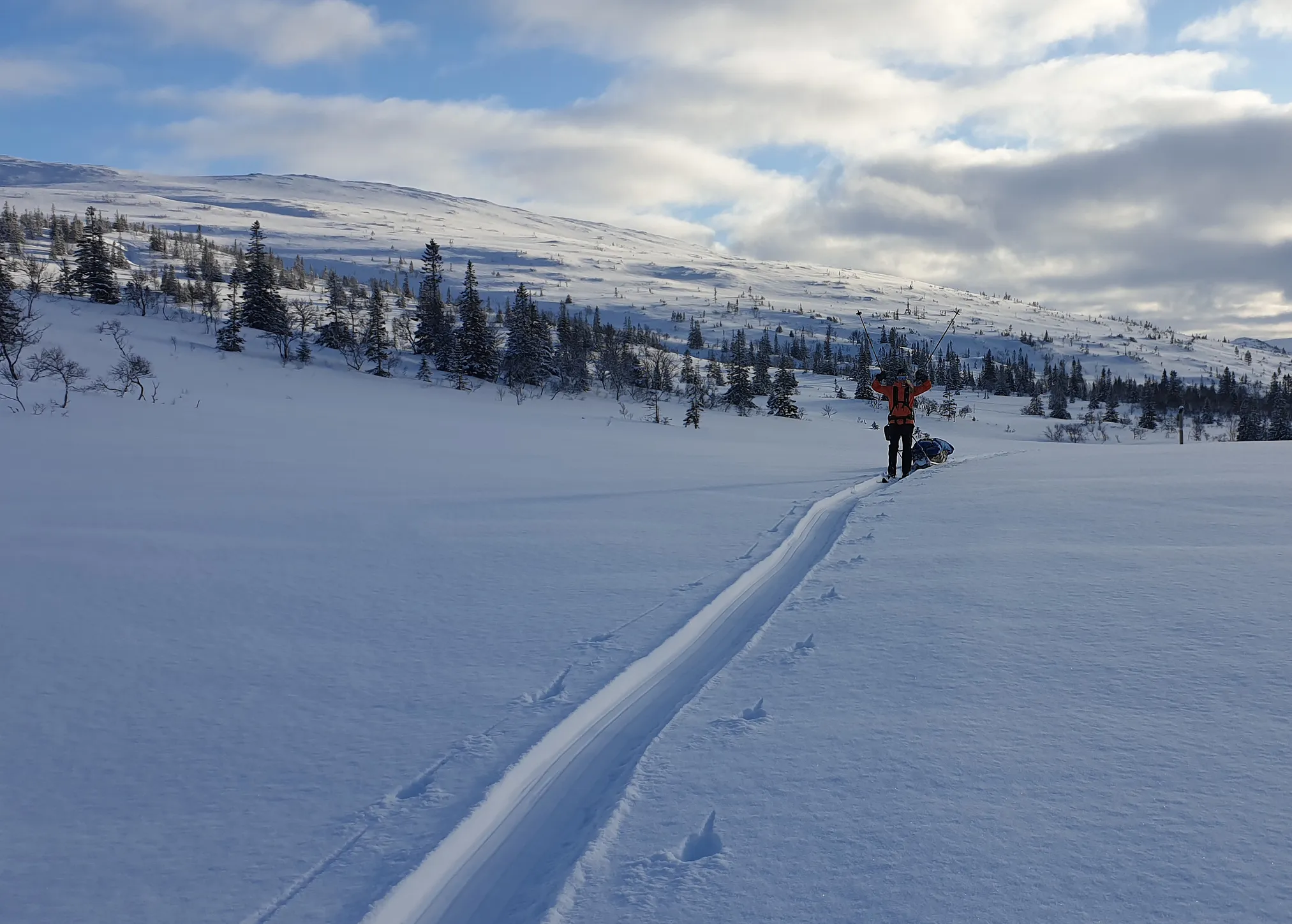 En person som går på langrenn over et stort snølandskap med fjell i bakgrunnen under en delvis skyet himmel.