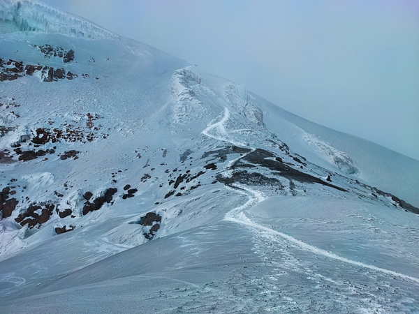 En snødekt fjellrygg med en svingete sti som leder oppover under en overskyet himmel.