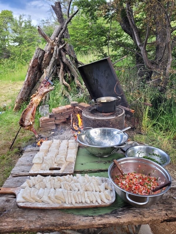 Et matlagingsområde utendørs har et trebord med oppskjærte poteter, boller med salat og kjøkkenredskaper. I bakgrunnen stekes kjøtt på spidd over et åpen ildsted med en gryte som koker over, i skogomgivelser.