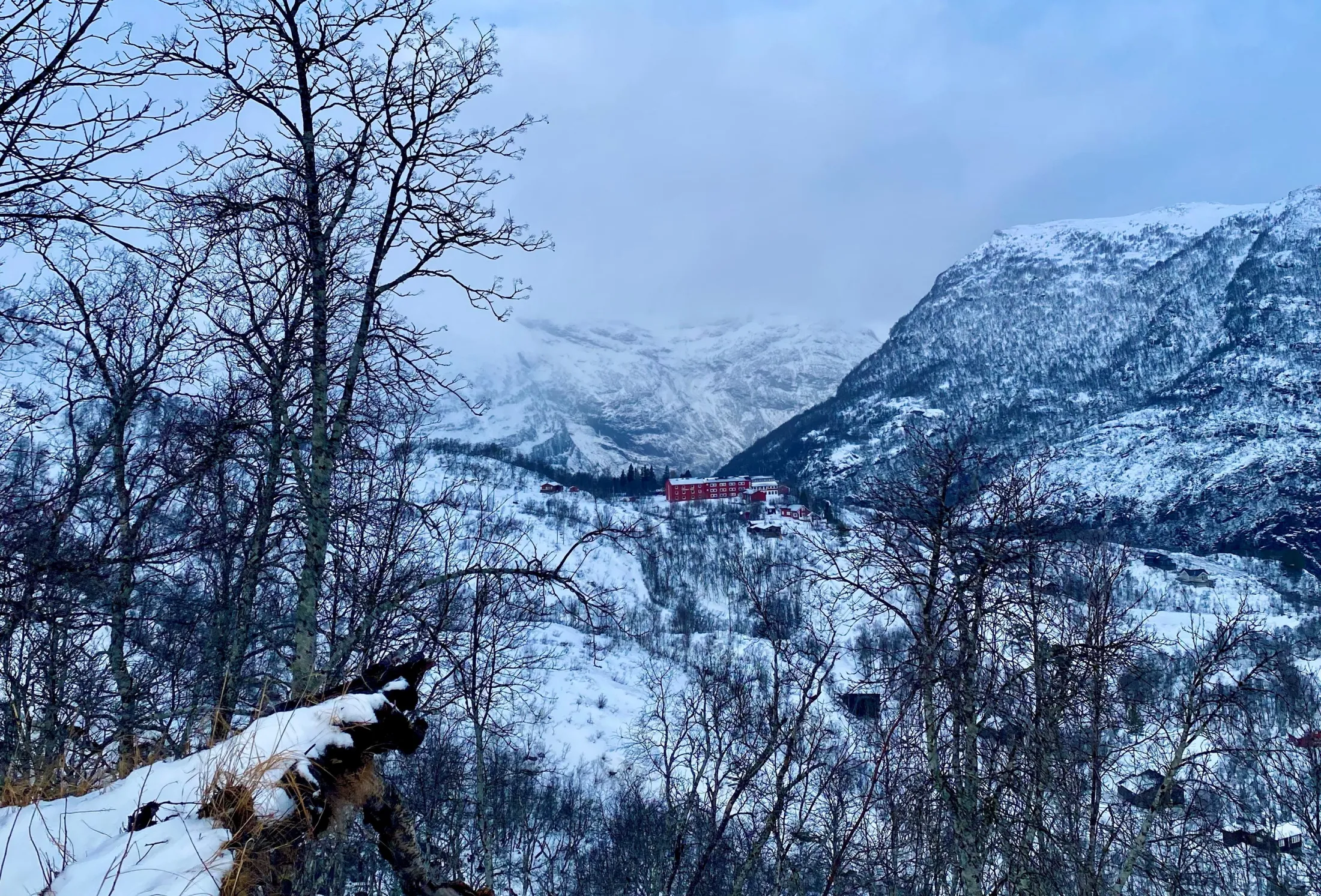 Røde hyttebygninger ligger i en snødekt helling omkranset av bladtomme trær og dramatiske snødekte fjell under en overskyet himmel.