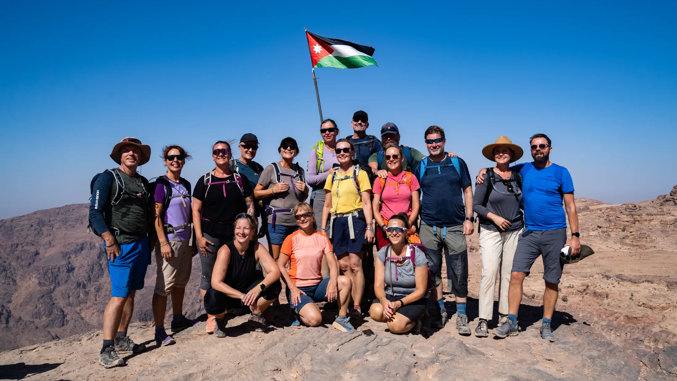 En gruppe turgåere poserer smilende på en fjelltopp under det jordanske flagget med utsikt over det vidstrakte landskapet.