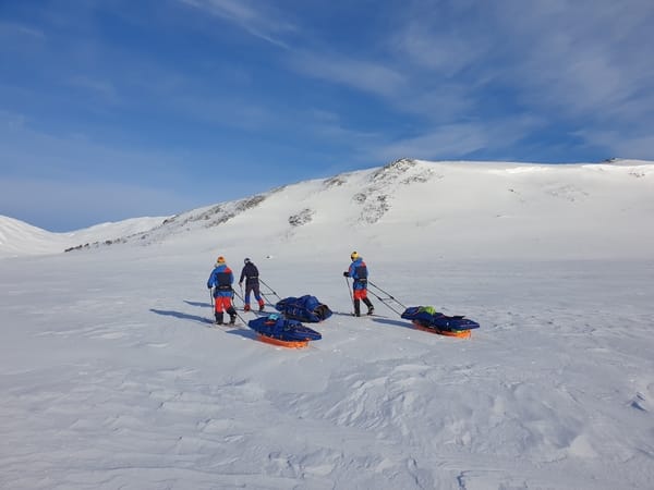 Tre personer med pulk i et snødekt fjelllandskap under en blå himmel.