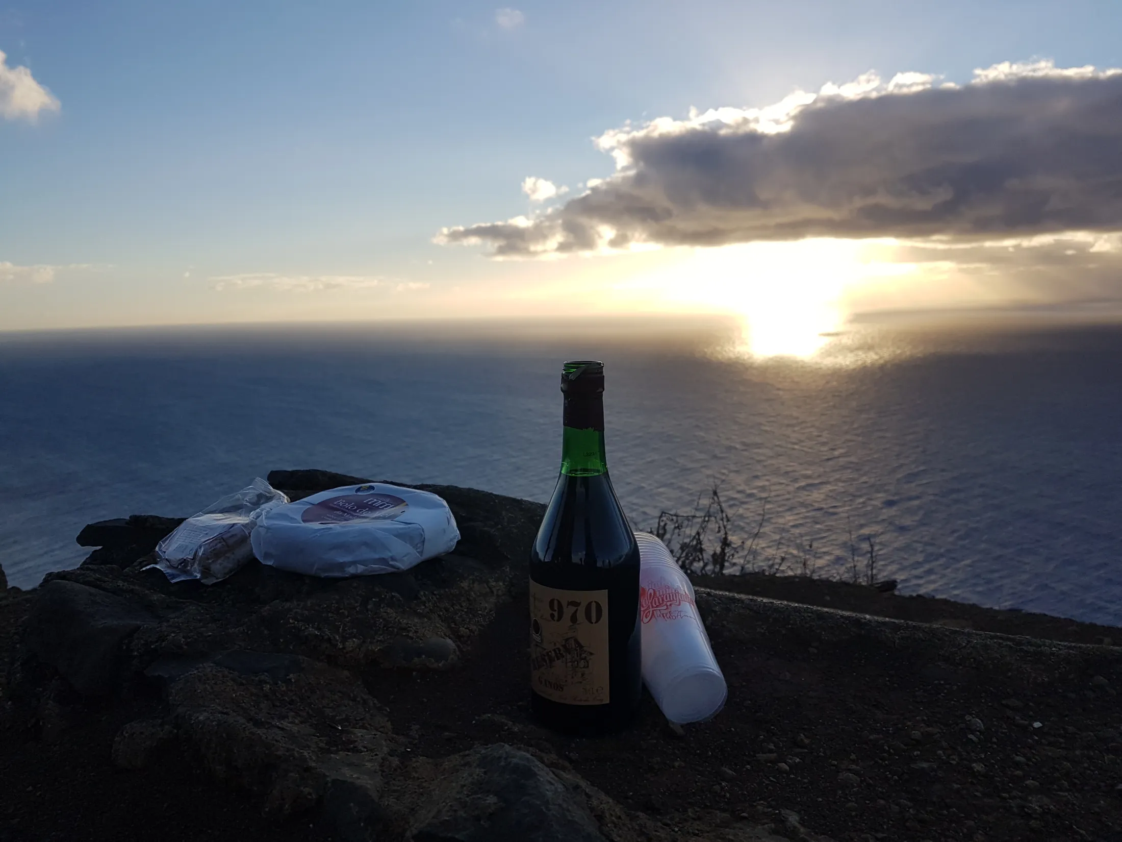 En vinflaske og mat på en stein med utsikt over havet ved solnedgang.