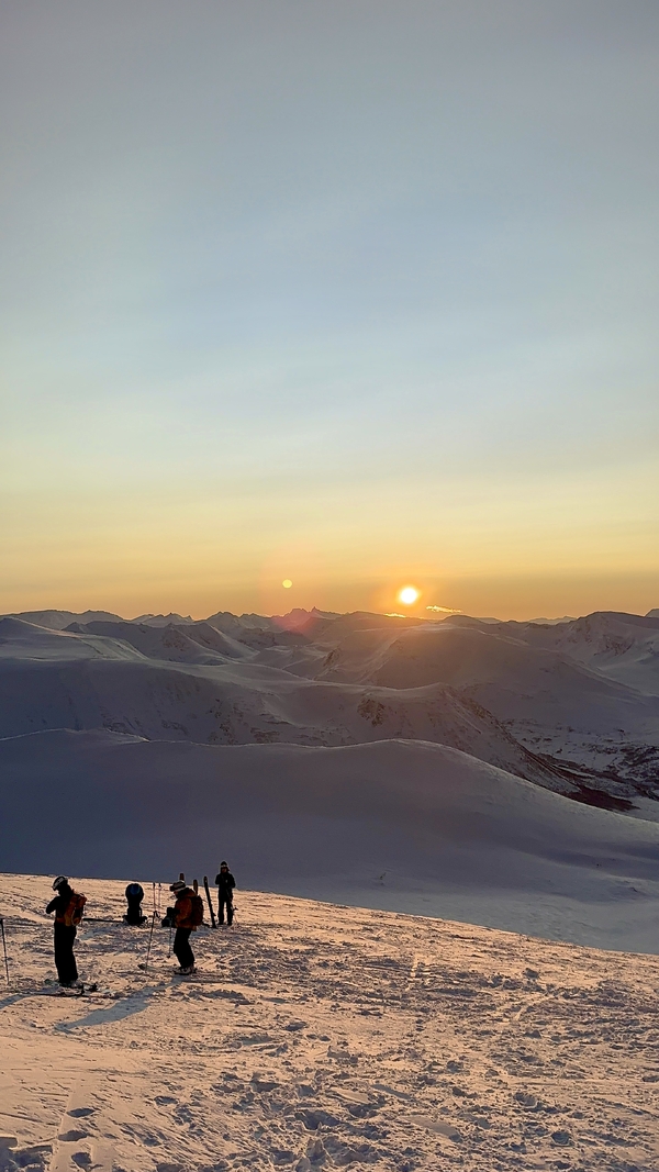 Skikjøpere står på en snødekt fjelltopp ved solnedgang og ser ut over et stort landskap av snødekte topper under en gyllen himmel.