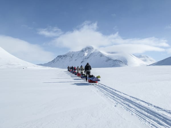 En rekke skiløpere som trekker pulker over en snødekt vidde mot fjerne fjell under en blå himmel.