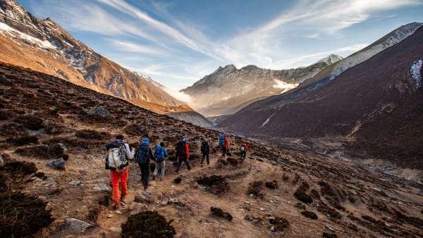 En gruppe turgåere med ryggsekker går på en rekke nedover en svingete jordsti gjennom en enorm, værbitte fjelldal mot kneisende fjelltopper opplyst av ettermiddagssolen.
