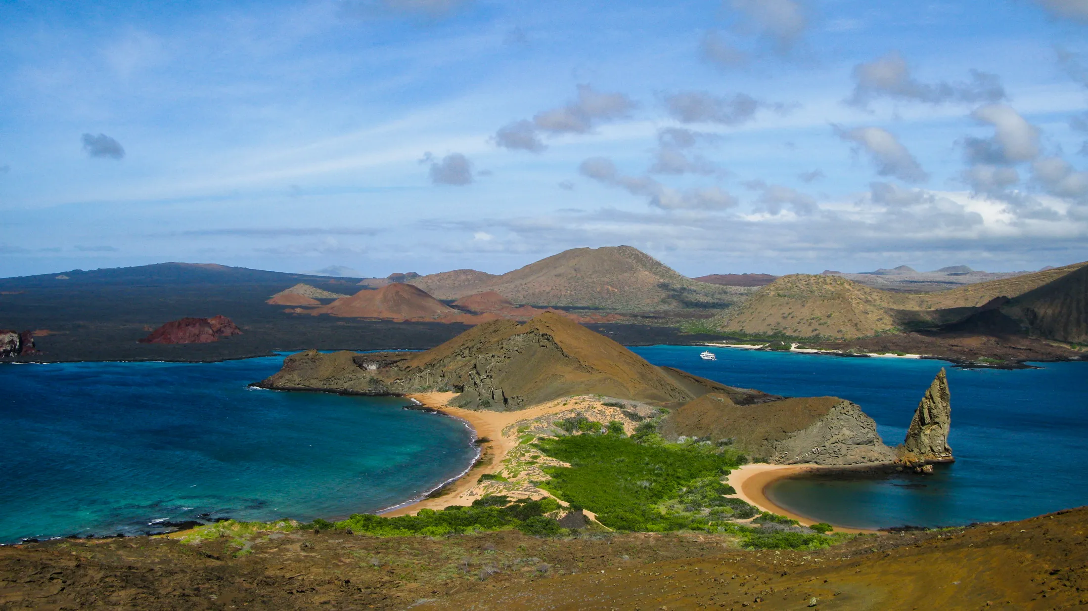 En opphøyd panoramautsikt over Bartolomé-øya på Galapagos, som viser Sullivan Bay, halvmåneformede sandstrender og den ikoniske Pinnacle Rock som reiser seg fra det dypblå havet.