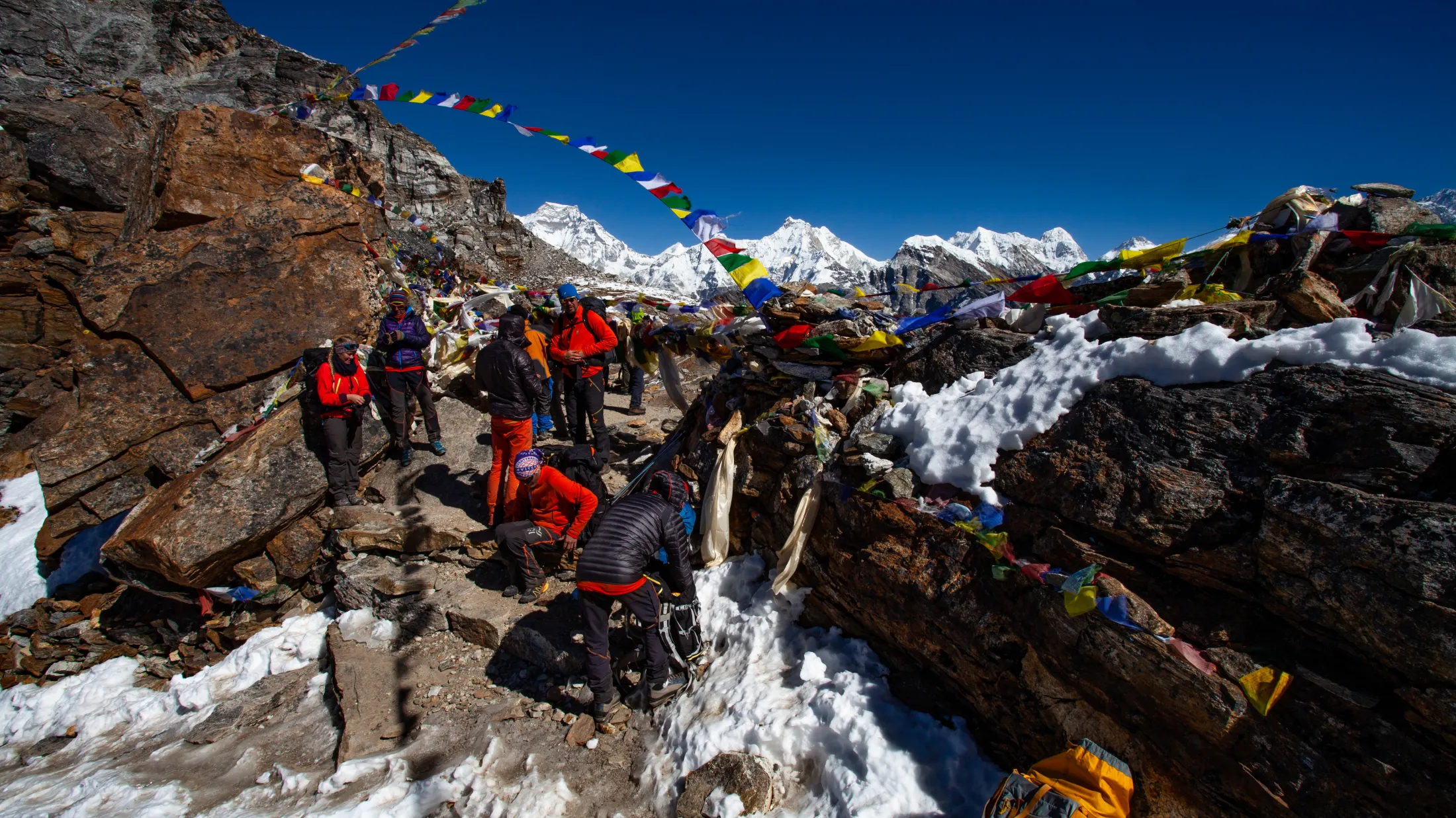 En gruppe fjellklatrere hviler på et snødekt og steinete fjellpass pyntet med fargerike buddhistiske bønneflagg, med en fantastisk panoramautsikt over snødekte Himalayatopper under en dyp blå himmel bak seg.