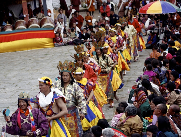 Deltakere i fargerike, tradisjonelle drakter og pyntede hodeplagg går i prosresjon gjennom en bakgård under en festival i Bhutan, omgitt av tilskuere.
