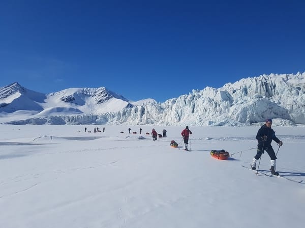 Skiløpere trekker ekspedisjonspulker over frossen sjøis foran en massiv, sprukken brevegg under en klar, blå himmel.