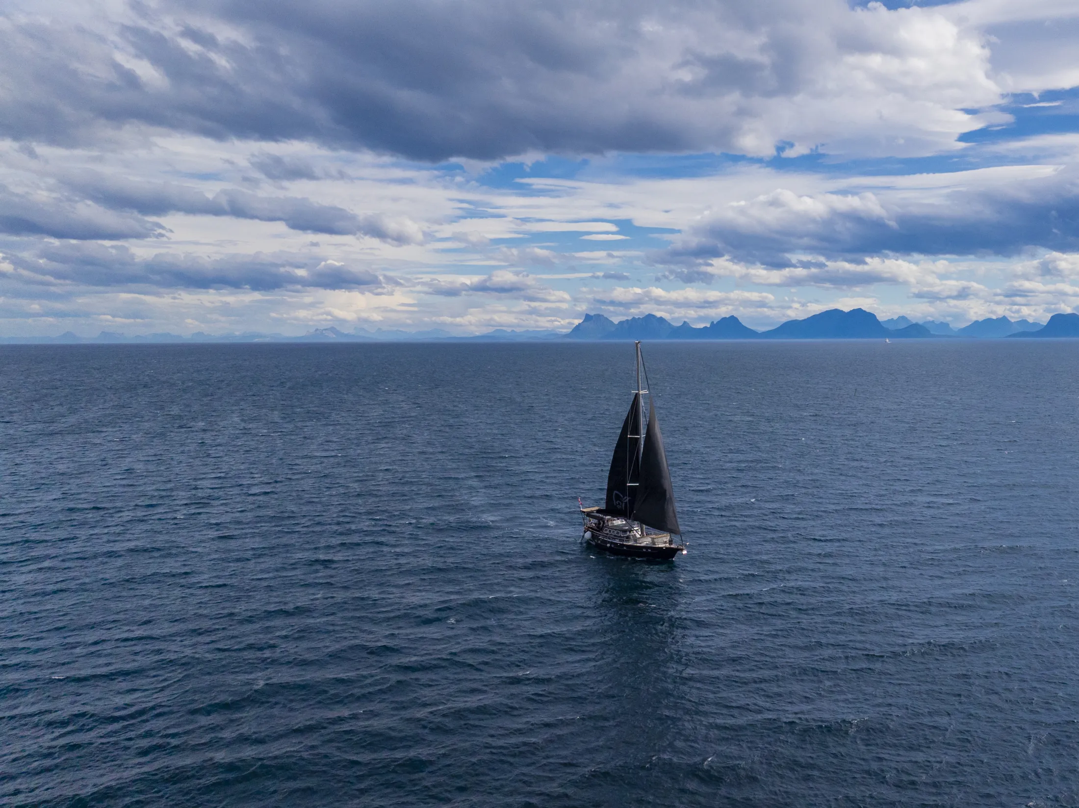 En seilbåt med mørke seil seiler på et rolig hav under en skyet himmel, med fjerne fjell i horisonten.