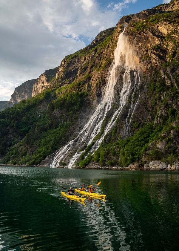 To gule kajakker med personer som padler i en fjord under en stor foss som fosser nedover et bratt, grønt fjell.